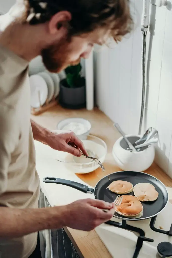 hombre de unos 24 años de edad cocinando pancakes de avena en una sarten en su casa